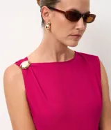 Vestido midi de sastrería color fucsia, sin mangas y con escote redondo. Presenta un adorno metálico dorado en uno de los hombros y una abertura lateral en la falda que aporta movimiento.