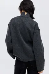 Buzo oversized de punto suave, color gris oscuro, con cuello alto acanalado y abertura trasera en V. Presenta costuras decorativas verticales y hombros caídos con mangas largas.