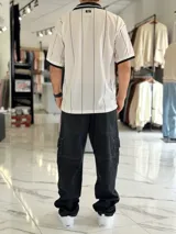Camiseta deportiva blanca de manga corta con rayas verticales negras, cuello polo en V de color negro con ribete blanco, y puños negros. Cuenta con estampado frontal con la palabra "TOKIO" en grande, además de detalles gráficos y texto en el pecho.