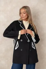 Tapado de paño negro con detalles de costuras y bordes en color blanco. Presenta un diseño de doble botonadura, solapas clásicas y corte amplio.