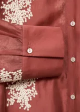 Camisa de manga larga en color terracota con diseño semitransparente. Presenta cuello camisero, cierre frontal con botones y bordados florales en color blanco distribuidos por toda la prenda.