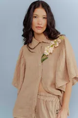 Camisa de lino color tostado, con cuello camisero y cierre frontal de botones. Se caracteriza por sus mangas cortas amplias y voluminosas que aportan un diseño fresco y liviano.