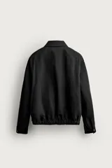 Cazadora negra de corte regular fit, confeccionada en tejido técnico con mezcla de algodón. Cuenta con cuello solapa, manga larga con puño abotonado, bolsillos de vivo en la cadera, pliegues laterales en la espalda y bajo elástico. Cierre frontal con cremallera.
