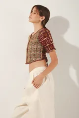 Saco corto estilo crop top con cuello redondo y mangas cortas. Confeccionado en tejido jacquard con lurex, presenta un estampado en tonos borgoña y dorado, y está ricamente bordado a mano con hilos y espejos (mostacillas). Cierre frontal con ganchos ocultos.