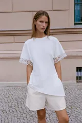 Camiseta negra de algodón con cuello redondo, manga corta y detalle de aplicación de encaje a tono en los puños. Presenta un diseño con bajo asimétrico y frunce lateral.