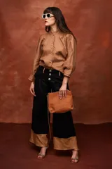 Pantalón ancho de tiro alto color beige, con pliegues frontales y botones visibles en la cintura. El ruedo del pantalón tiene un detalle de terminación en cuero color caramelo.