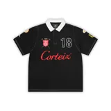 Remera polo de manga corta de la marca Corteiz, color negro, con cuello blanco y cierre de botones oculto. Presenta detalles bordados y estampados, incluyendo el texto 'Corteiz' en rojo en el pecho, el número '18' y diversos parches en las mangas.
