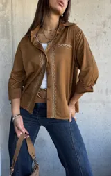Camisa de gamuza color camel, con cuello clásico, cierre frontal de botones y detalle de bordado geométrico en color crema a lo largo de la botonadura y en el bolsillo frontal. Presenta mangas largas con puños doblados.