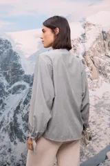 Camisa de tela afelpada color verde grisáceo, con cuello con lazo anudado, mangas largas abullonadas con detalles de lazos en los puños y cierre frontal con botones.
