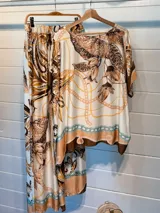 Blusa de satén con estampado de cadenas y hojas en tonos blanco, marrón y negro. Tiene corte amplio, mangas cortas y cuello redondo. Se ajusta a la cintura con un cinturón elástico negro con hebilla dorada.