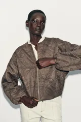 Chaqueta bomber color marrón con cuello redondo, manga larga con puño elástico y cierre frontal de cremallera metálica. Presenta un diseño de bordados florales en relieve por toda la prenda y bolsillos delanteros de vivo.