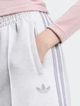 Pantalón deportivo Adidas modelo Spacer, color gris melange, de corte holgado y pierna ancha con pliegues frontales. Cuenta con cintura elástica, bolsillos laterales y las clásicas tiras laterales de la marca en tono gris oscuro, además del logo del Trifolio bordado.