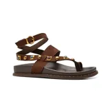 Sandalias planas Korium Adhara de cuero color marrón oscuro con tiras de gamuza en tono tostado. Presentan diseño de tira al dedo, tachas doradas y abalorios de piedras incrustadas, y tira ajustable al tobillo con hebilla metálica.