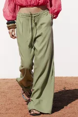 Pantalón ancho de tiro alto color beige claro, con cintura elástica, cordón ajustable y pinzas delanteras. Presenta bolsillos frontales y bolsillos de plastrón en la espalda. Incluye un cinturón del mismo tejido con apliques metálicos.