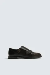 Zapato de vestir estilo monk strap fabricado en piel color marrón oscuro. Presenta ajuste mediante dos hebillas laterales metálicas, detalle de flecos en el empeine y puntera con perforaciones decorativas tipo brogue.