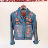 Campera de jean azul con diseño pintado a mano de estilo abstracto, con detalles en tonos turquesa, rosa y dorado. Cuenta con bolsillos en el pecho y cierre frontal con botones metálicos.