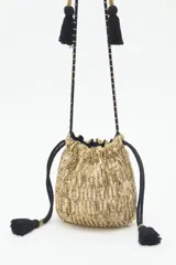 Cartera tipo saco de tamaño pequeño, confeccionada con tejido brillante en color dorado. Presenta cierre mediante cordones ajustables con borlas negras en los extremos y una correa larga de hombro con detalles decorativos y borlas adicionales.
