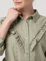 Blusa de algodón orgánico color verde oliva, con cuello camisero, cierre frontal de botones y mangas largas. Presenta detalles de volantes bordados en el frente y bordados calados en toda la prenda.