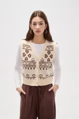 Chaleco tejido de punto con diseño jacquard de corazones y motivos geométricos en tonos crema y marrón. Presenta escote en V, cierre frontal con botones brillantes y terminaciones acanaladas.