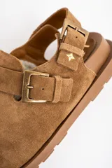 Zueco de gamuza color camel, con interior forrado en corderito, tira ajustable en el talón y suela de goma plana.