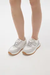 Zapatilla deportiva de diseño urbano, combinando paneles de tela blanca y gamuza sintética en tono gris claro. Cuenta con cordones grises, suela de plataforma con entresuela blanca y base dentada en color caramelo.