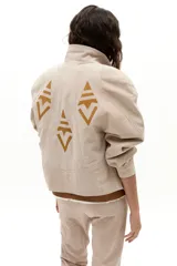 Chaqueta oversized de gamuza de cabra color arena, con apliques tribales en cuero de oveja color mostaza. Presenta hombros exagerados, cuello camisero redondeado, bolsillos frontales y botones a presión forrados. Cuenta con forro capitoneado y un bolsillo interno.