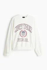 Buzo de algodón con cuello redondo, hombros caídos y puños acanalados. Presenta un estampado frontal con estilo universitario que incluye el texto 'NEW YORK PHYS.ED.' y un emblema central en tonos rojo y azul.