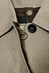 Trench coat largo color verde oliva/beige, con cuello solapa, manga larga ajustable con trabilla y botón, bolsillos delanteros y cierre frontal con botones. El forro interior visible en el cuello es de tela estampada a cuadros en tonos marrones.