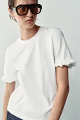 Camiseta blanca de algodón con cuello redondo y manga corta, con detalle de aplicaciones de flores en relieve en los dobladillos de las mangas.