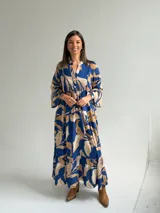Vestido largo azul con estampado de hojas en tonos beige y blanco, escote en V con botones frontales, mangas largas amplias y diseño escalonado.