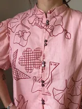 Camisa de popelina de algodón con cuello mao, botones frontales agrupados en pares y mangas cortas abullonadas tipo balloon. Cuenta con un diseño estampado de patrón geométrico y floral sobre fondo negro.