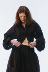 Camisa oversize negra de algodón liviano con textura calada, cuello camisero, botones frontales, mangas largas voluminosas y tiras para anudar en la espalda.
