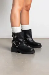 Botas de caña baja en cuero sintético color marrón, con diseño estilo biker que incluye dos correas con hebillas metálicas y una argolla decorativa en el lateral. Poseen taco bajo y suela de goma negra.