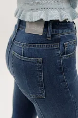 Jean de denim azul de corte acampanado, con costuras frontales en relieve tipo alforza y terminación desmechada en el bajo.