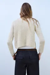 Chaqueta de punto de hilo metalizado color beige con cuello redondo, manga larga y cierre frontal con un botón dorado.