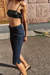 Jeans de tiro medio en denim azul oscuro, con diseño capri de pernera entallada. Presenta cierre frontal con cremallera y botón, y un cinturón integrado con hebilla metálica en la cintura.