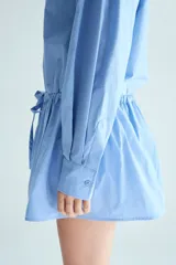 Vestido camisero corto de popelina de algodón en color celeste, con cuello clásico, cierre frontal con botones, mangas largas con puños sencillos y cordón ajustable en la cintura.