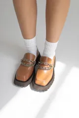 Mocasín de cuero color suela con punta cuadrada y suela track de goma oscura. Presenta un detalle de doble cadena metálica plateada sobre el empeine.