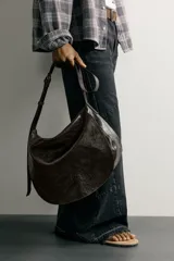 Bolso bandolera de piel color marrón oscuro, con diseño flexible y solapa frontal. Cuenta con asa larga ajustable y compartimento principal con bolsillo interior con cierre.