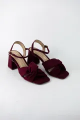 Sandalias de tacón cuadrado color bordó, confeccionadas en material textil simil gamuza. Presentan un diseño de tiras cruzadas con nudo frontal, puntera cuadrada y cierre de pulsera con hebilla metálica en el tobillo.