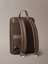 Mochila de hombre con silueta slim y cuadrada, confeccionada en material sintético con textura microgranulada (micro pebble). Color marrón oscuro, con asa de mano superior, correas acolchadas para el hombro, cierre de cremallera doble y bolsillo exterior frontal con cierre.