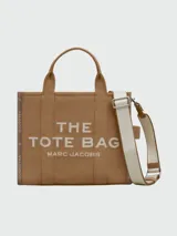 Bolso tipo tote de lona color negro con letras blancas bordadas en el frente que dicen 'THE TOTE BAG MARC JACOBS'. Cuenta con asas superiores cortas y una correa de hombro ajustable y extraíble.
