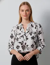 Blusa de gasa con estampado floral en tonos blanco y negro, cuello a la base con escote en V y cierre frontal parcial con botones. Presenta mangas tres cuartos con puños rectos.