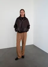 Chaqueta de cuero de napa marrón oscuro, de diseño oversize, con capucha ajustable, cierre frontal de cremallera, bolsillos laterales y bajo ajustable con cordón.