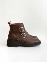 Botas cortas de cuero color marrón con suela track negra. Presentan dos correas ajustables con hebillas metálicas plateadas en el empeine y cierre lateral interno.