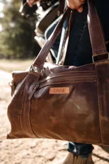 Bolso de cuero hecho a mano en color tostado, con diseño amplio y funcional. Presenta cierre superior, bolsillo frontal con cremallera, asas de mano reforzadas y una correa larga ajustable y desmontable.