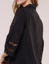 Camisa negra de poliéster con cuello camisero, escote en V y cierre frontal de botones. Cuenta con mangas anchas de corte acampanado y detalles de bordado suizo calado en las mangas y en el ruedo inferior.