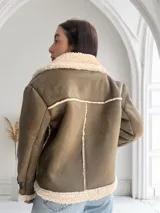 Chaqueta verde militar de gamuza sintética con interior de piel sintética color crema. Tiene cierre frontal con cremallera, cuello con solapas grandes y cinturón con hebilla en los puños.