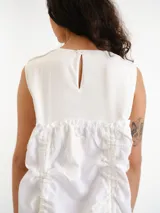 Vestido midi blanco sin mangas, con cuello redondo y diseño holgado. Presenta un cordón ajustable fruncido en el centro de la parte frontal y parches circulares sutiles en la tela.