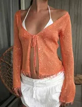 Cardigan tejido liviano de color coral con detalles de lentejuelas, terminaciones desflecadas y mangas largas acampanadas. Cuenta con escote en V y lazo delantero regulable.